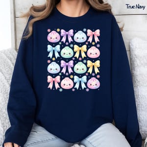 May include: A true navy crewneck sweatshirt featuring a repeating pattern of pastel-colored kawaii-style dumplings with smiling faces, interspersed with colorful bows and small flower icons. The text True Navy is printed in the top right corner.