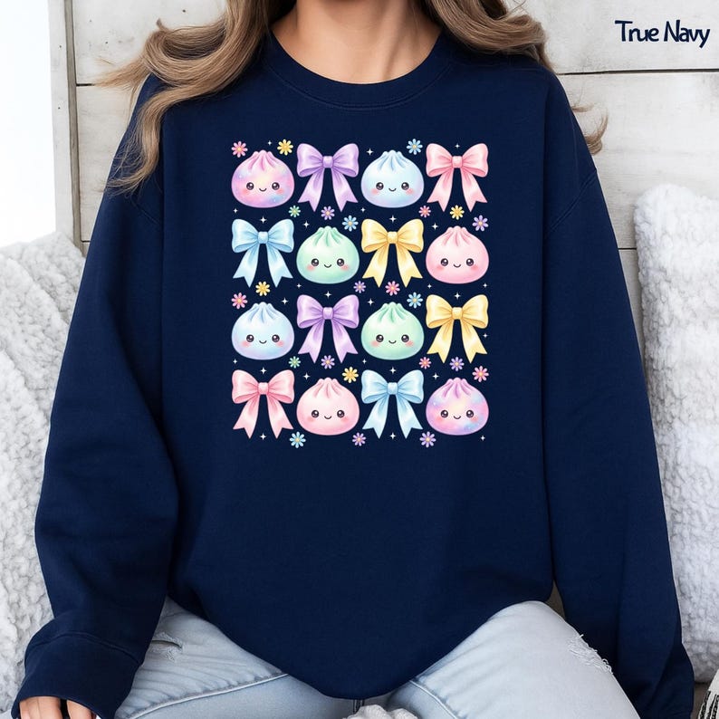 May include: A true navy crewneck sweatshirt featuring a repeating pattern of pastel-colored kawaii-style dumplings with smiling faces, interspersed with colorful bows and small flower icons. The text True Navy is printed in the top right corner.