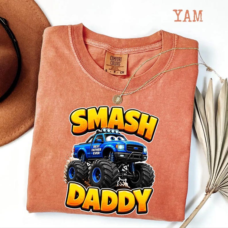 Może przedstawiać: Koralowa koszulka Comfort Colors z niebieską grafiką monster trucka i słowami "SMASH DADDY" w kolorze ż&oacute;łtym i pomarańczowym. Na ciężar&oacute;wce widnieje napis "BEST FATHER EVER". Widoczny jest r&oacute;wnież złoty naszyjnik i brązowy kapelusz.