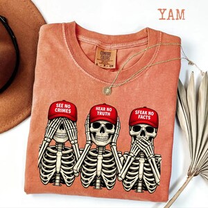 May include: Coral-colored Comfort Colors t-shirt featuring three skeleton figures in the 'see no evil, hear no evil, speak no evil' pose. Each skeleton wears a red cap with a different phrase. A gold necklace and a brown hat are also visible.
