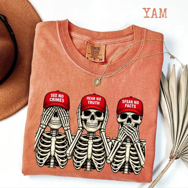 May include: Coral-colored Comfort Colors t-shirt featuring three skeleton figures in the 'see no evil, hear no evil, speak no evil' pose. Each skeleton wears a red cap with a different phrase. A gold necklace and a brown hat are also visible.