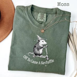 Könnte beinhalten: Ein moosgrünes T-Shirt mit einer Grafik eines Opossums, das einen Zaubererhut trägt und Roller fährt. Der Text "Off To Cause A Kerfuffle" ist unter der Grafik aufgedruckt. Eine goldene Halskette liegt auf dem Shirt.