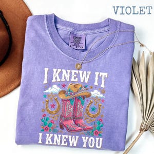 May include: A violet short-sleeve t-shirt with a graphic print featuring a cowboy hat, two pink cowboy boots, horseshoes, and cacti. The text reads I KNEW IT I KNEW YOU. The shirt has a crew neckline and a Comfort Colors brand label at the neck.