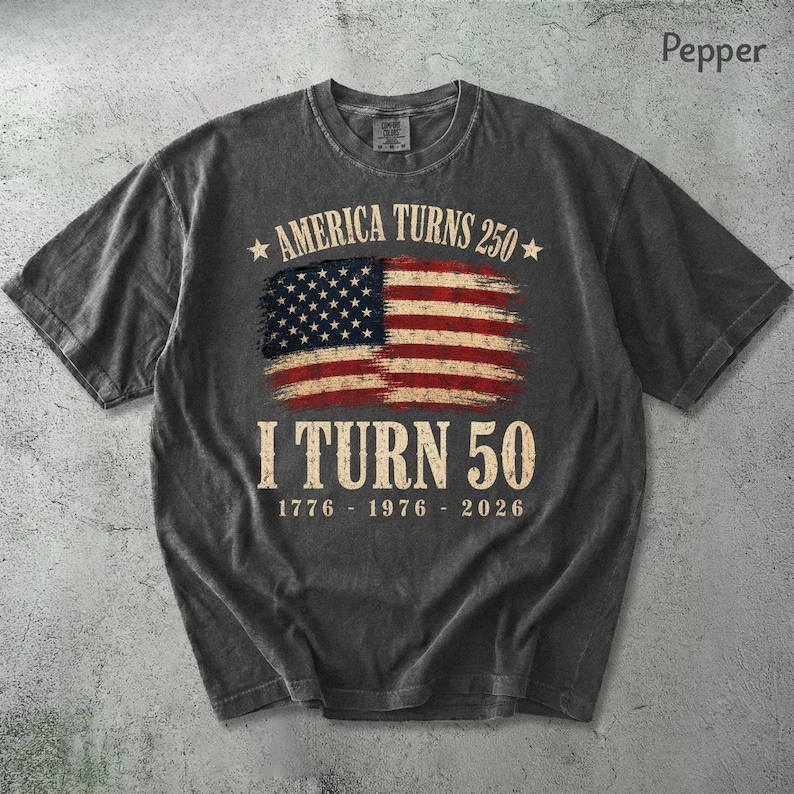 May include: A dark gray t-shirt with a distressed American flag graphic. The text reads "AMERICA TURNS 250" above the flag and "I TURN 50" below, with the years 1776-1976-2026. The shirt has a crew neck and short sleeves.