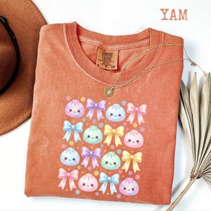 May include: A terracotta-colored short-sleeve t-shirt featuring a graphic print of various pastel-colored bows and smiling dumpling characters. The shirt has a crew neckline and a Comfort Colors brand label on the inside collar area.