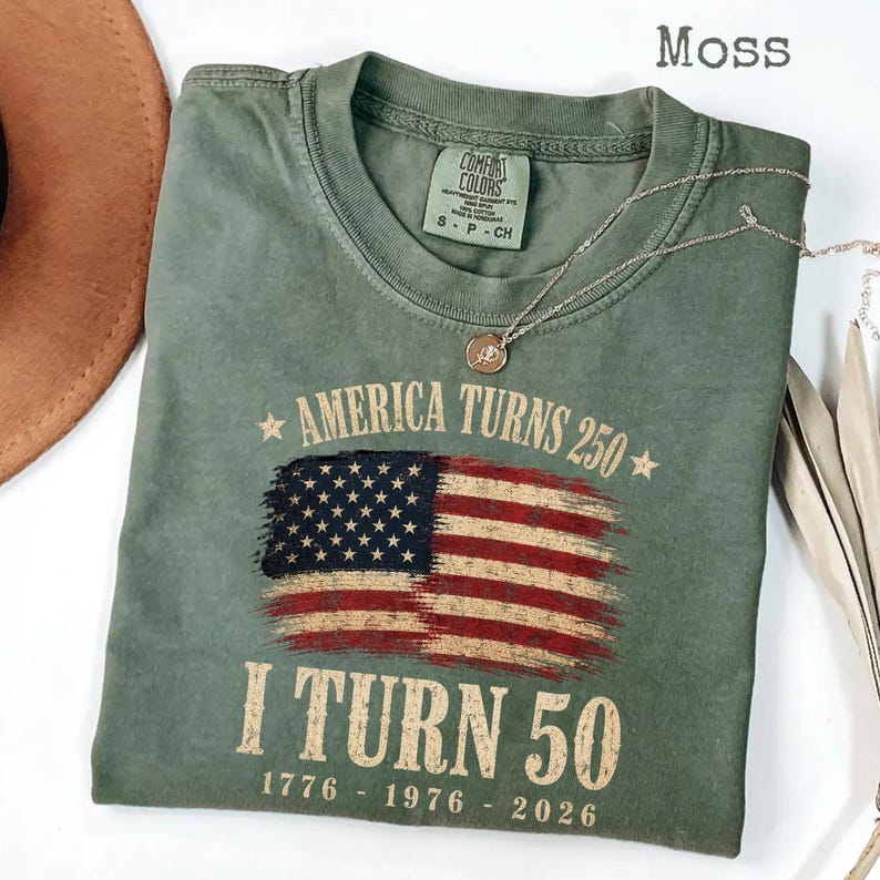May include: Moss green t-shirt with the text "America Turns 250" and "I Turn 50" printed above and below a distressed American flag graphic. The shirt has a crew neck and short sleeves. A gold necklace with a pendant is draped on the shirt.