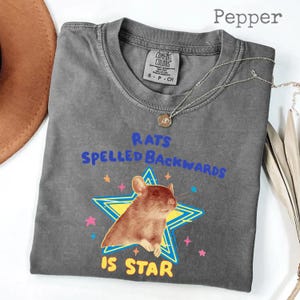 May include: Pepper-colored Comfort Colors t-shirt with the text "RATS SPELLED BACKWARDS IS STAR" and a graphic of a rat inside a star. The shirt has a crew neck and short sleeves. A silver necklace with a pendant is draped across the shirt.