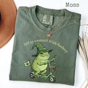 Könnte beinhalten: Moosgrünes T-Shirt mit einer Grafik einer Froschfigur, die einen Leprechaun-Hut trägt und einen Roller fährt, mit einem vierblättrigen Kleeblatt. Der Text "Off to commit Irish foolery" ist darüber. Eine goldene Halskette liegt auf dem Shirt.