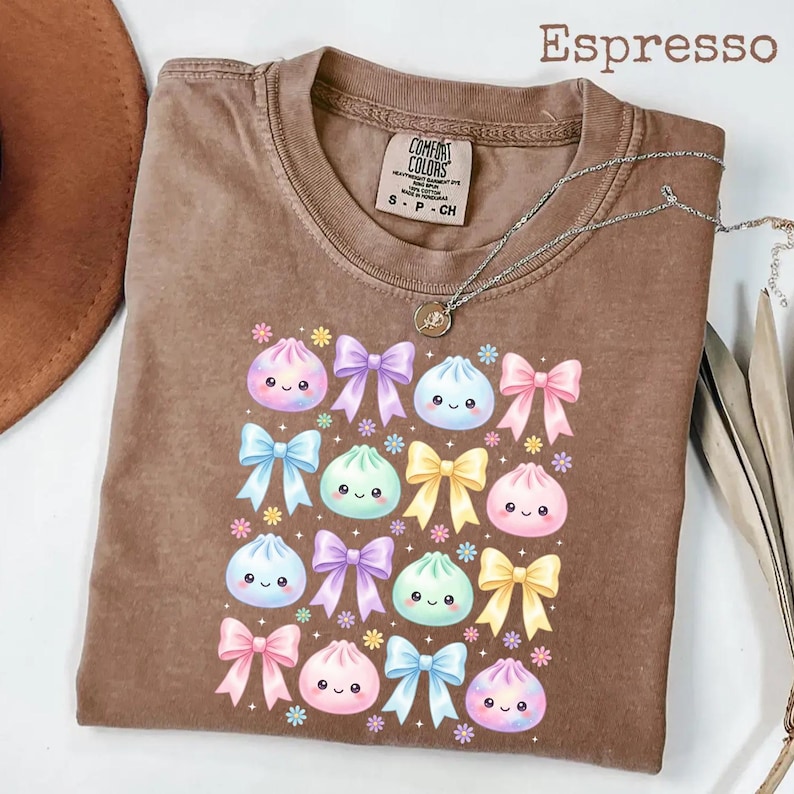 May include: An espresso-colored t-shirt featuring a graphic print of various pastel-colored dumplings with smiling faces interspersed with colorful bows and small flowers. The shirt has a crew neckline and a Comfort Colors brand label inside.