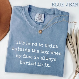 Puede incluir: Camiseta azul vaquero con el texto "it's hard to think outside the box when my face is always buried in it." Un collar dorado con un colgante está en la camiseta. La camiseta tiene una etiqueta Comfort Colors.