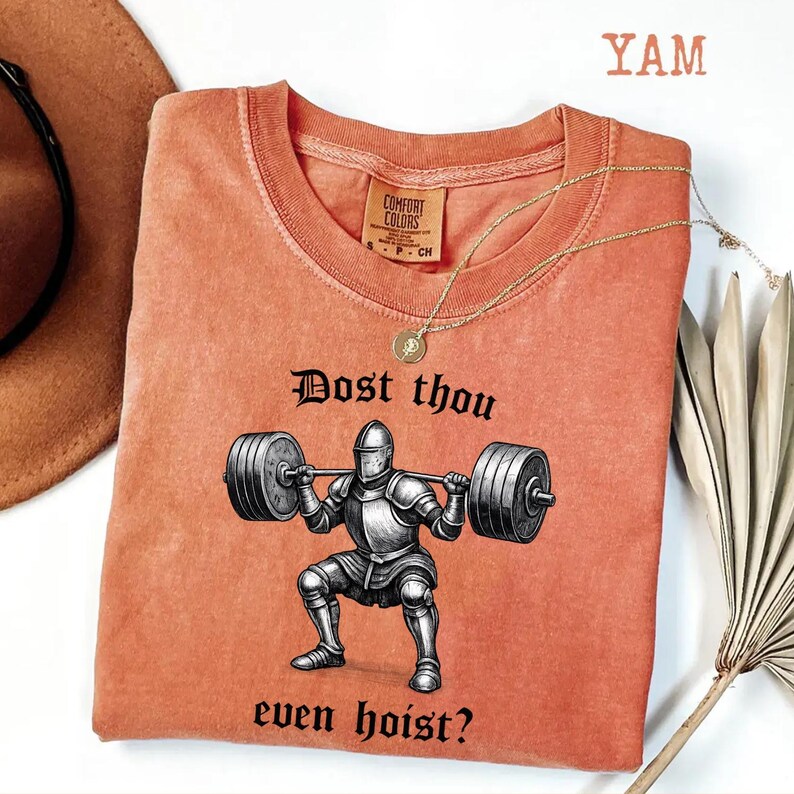 May include: Coral-colored t-shirt with a graphic of a knight lifting weights, and the text "Dost thou even hoist?" A gold necklace and a Comfort Colors tag are visible. The shirt is a casual wear item.
