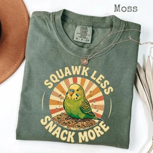 Könnte beinhalten: Moosgrünes T-Shirt mit einer Grafik eines grünen Wellensittichs und dem Text "Squawk Less Snack More". Der Wellensittich sitzt auf einem Haufen Samen, vor einem Retro-Sonnenstrahl-Design. Eine silberne Halskette liegt über dem Shirt.