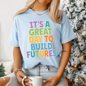 Puede incluir: Camiseta azul claro con la frase "IT'S A GREAT DAY TO BUILD FUTURES" en letras de colores. La camiseta es de estilo casual, perfecta para el uso diario. El texto es en morado, verde, naranja, azul y amarillo.