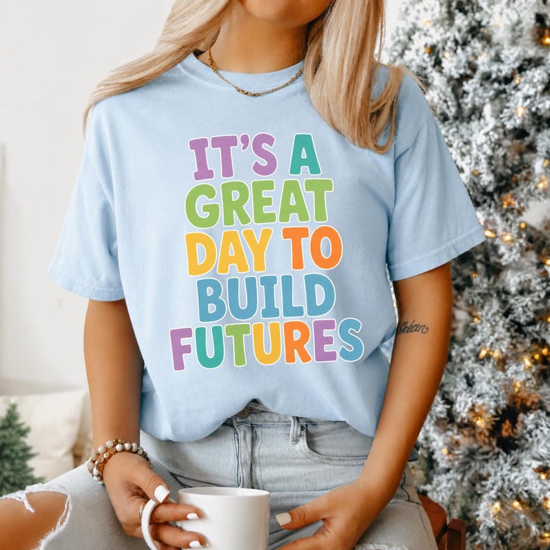 Puede incluir: Camiseta azul claro con la frase "IT'S A GREAT DAY TO BUILD FUTURES" en letras de colores. La camiseta es de estilo casual, perfecta para el uso diario. El texto es en morado, verde, naranja, azul y amarillo.