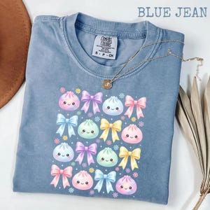 May include: A blue jean colored t-shirt featuring a graphic print of various pastel-colored bows and smiling dumpling characters. The shirt has a crew neckline and a Comfort Colors brand label on the inside collar. A gold chain necklace rests on top.