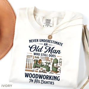 Op de afbeelding: Ivoor kleurig t-shirt met de tekst "Never Underestimate an Old Man Who Still Does Woodworking In His Eighties." Het ontwerp bevat houtbewerkingsgereedschap en machines. Een gouden ketting en een hoed zijn gedeeltelijk zichtbaar.