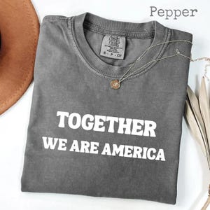 Peut inclure: Un t-shirt gris Comfort Colors avec le texte "TOGETHER WE ARE AMERICA" en lettres blanches. Un collier argenté avec un pendentif repose sur le t-shirt. Le t-shirt est plié et présenté avec un chapeau marron.
