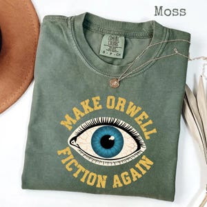 May include: A moss green t-shirt with the slogan "MAKE ORWELL FICTION AGAIN" in yellow. The design includes a blue eye. A silver necklace with a pendant is draped on the shirt. The shirt is a Comfort Colors brand.
