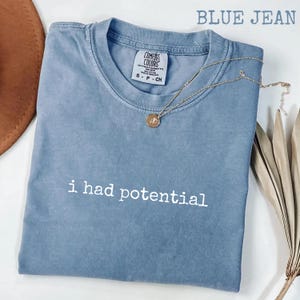 May include: A blue jean Comfort Colors t-shirt with the phrase "i had potential" in white lettering. A gold necklace with a pendant rests on the shirt. The shirt is folded and placed on a white surface.