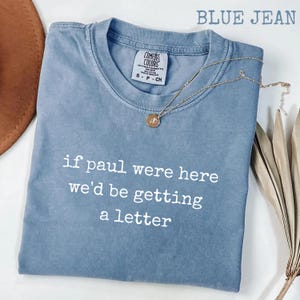 May include: Blue jean Comfort Colors t-shirt with the text "if paul were here we'd be getting a letter" in white. A gold necklace with a pendant rests on the shirt. The shirt is folded on a white surface.
