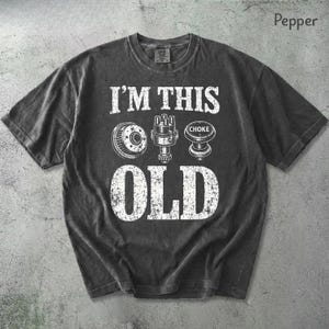 May include: A pepper-coloured t-shirt featuring the text "I'M THIS OLD" in white, accompanied by illustrations of car components. The shirt has a vintage, distressed appearance.