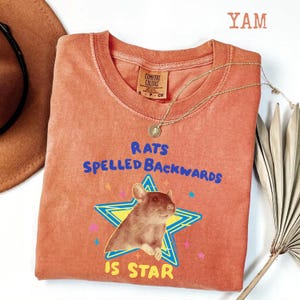 May include: A coral-colored t-shirt with the text "RATS SPELLED BACKWARDS IS STAR" in blue and yellow. The graphic features a rat inside a star. A gold necklace and a brown hat are also visible.