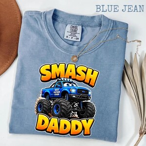 Może przedstawiać: Niebieska koszulka Comfort Colors z nadrukiem niebieskiego monster trucka z napisem "SMASH DADDY" w kolorze ż&oacute;łtym i pomarańczowym. Widoczny jest r&oacute;wnież złoty naszyjnik.