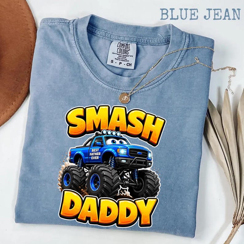Może przedstawiać: Niebieska koszulka Comfort Colors z nadrukiem niebieskiego monster trucka z napisem "SMASH DADDY" w kolorze ż&oacute;łtym i pomarańczowym. Widoczny jest r&oacute;wnież złoty naszyjnik.