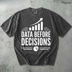 K&ouml;nnte beinhalten: Ein anthrazitfarbenes Kurzarm-T-Shirt mit wei&szlig;em Grafikdruck. Das Design zeigt ein nach oben gerichtetes Balkendiagramm, den Text "DATA BEFORE DECISIONS" in fetten Gro&szlig;buchstaben und ein kreisf&ouml;rmiges Tortendiagramm-Symbol unter dem Text.