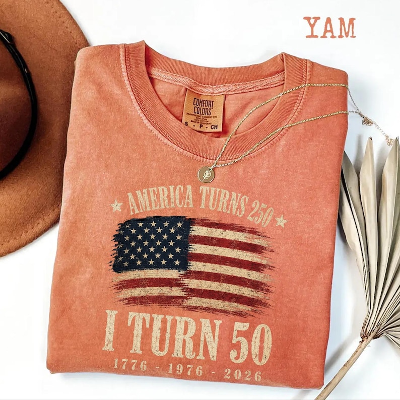May include: A coral-colored t-shirt with the American flag design. The shirt has the text "America Turns 250" and "I Turn 50" with the years 1776, 1976, and 2026. A gold necklace and a brown hat are also visible.