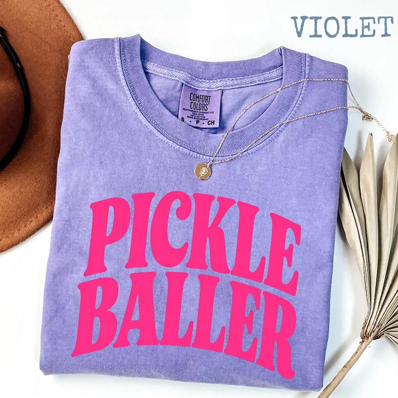 Op de afbeelding: Een violetkleurig T-shirt met de woorden "PICKLE BALLER" in felroze, gebogen over de voorkant. Het shirt heeft een ronde hals en een klein label bij de kraag. Een gouden ketting met een hanger is over het shirt gedrapeerd.