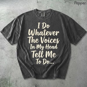 May include: A charcoal grey short-sleeve t-shirt featuring the cream-coloured text I Do Whatever The Voices In My Head Tell Me To Do... printed in a distressed, serif font across the centre of the chest. The garment has a crew neckline.