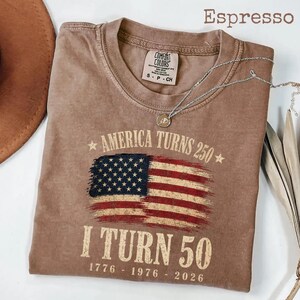 May include: Espresso-colored t-shirt with the text "America Turns 250" and "I Turn 50" printed on the front. The design includes an American flag graphic. A silver necklace with a pendant is draped on the shirt.