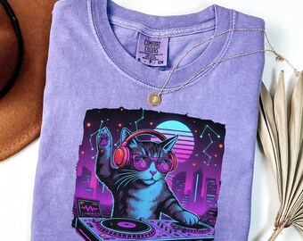 May include: Violet Comfort Colors t-shirt featuring a graphic of a cat DJ wearing headphones and sunglasses, with a cityscape and a retro sunset design. The shirt has a crew neck and short sleeves. A gold necklace is draped across the shirt.