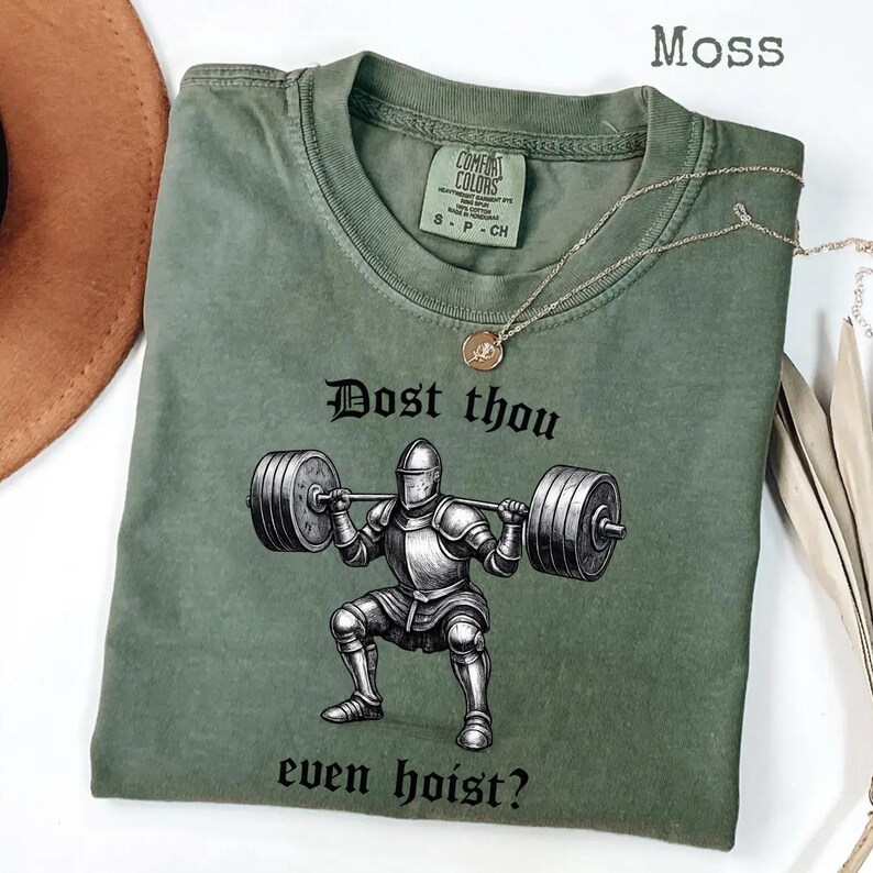 May include: Moss green t-shirt with the text "Dost thou even hoist?" and a graphic of a knight lifting weights. The shirt has a crew neck and short sleeves. A gold necklace is draped across the shirt.