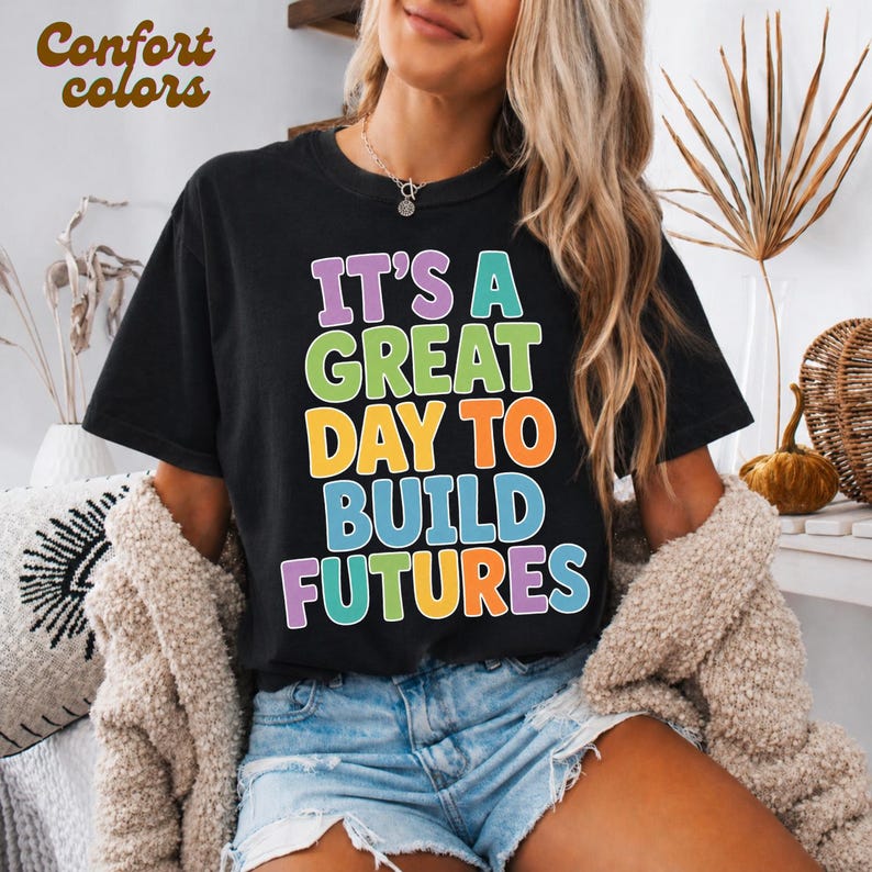 Puede incluir: Camiseta negra con la frase colorida "IT'S A GREAT DAY TO BUILD FUTURES". Las palabras "Comfort Colors" est&aacute;n en la esquina superior izquierda. La camiseta se combina con pantalones cortos de mezclilla y una rebeca crema.