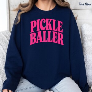Op de afbeelding: Een marineblauw sweatshirt met de woorden "PICKLE BALLER" in helder roze, vette retro-letters. De tekst "True Navy" is zichtbaar in de rechterbovenhoek. Het sweatshirt is een casual stijl.