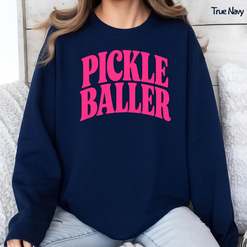 Op de afbeelding: Een marineblauw sweatshirt met de woorden "PICKLE BALLER" in helder roze, vette retro-letters. De tekst "True Navy" is zichtbaar in de rechterbovenhoek. Het sweatshirt is een casual stijl.