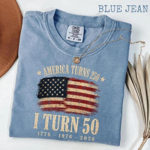 May include: A blue jean Comfort Colors t-shirt with the text "America Turns 250" and "I Turn 50" printed in gold. The shirt features a distressed American flag graphic. A gold necklace with a pendant is draped across the shirt.