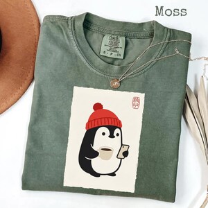 May include: A moss green short-sleeve t-shirt featuring a graphic of a penguin wearing a red beanie, holding a coffee mug and a smartphone. The shirt has a Comfort Colors label at the neck and the text Moss is printed in the upper right corner.