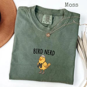 May include: A moss green t-shirt with the words "BIRD NERD" printed in black. The shirt features a cartoon bird wearing a hat and binoculars. A silver necklace with a small pendant is draped across the shirt.