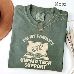 May include: Moss green t-shirt with the text "I'M MY FAMILY'S UNPAID TECH SUPPORT" and a laptop graphic. The shirt has a crew neck and short sleeves. A gold necklace with a pendant is draped across the shirt.