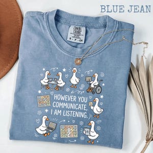 Op de afbeelding: Blauw Comfort Colors T-shirt met een afbeelding van witte cartoon ganzen die verschillende communicatiemethoden gebruiken. De tekst luidt: "HOWEVER YOU COMMUNICATE, I AM LISTENING." Een gouden ketting met hanger ligt op het shirt.