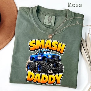 Może przedstawiać: Zielona koszulka w kolorze mchu z napisem "SMASH DADDY" w ż&oacute;łtych i pomarańczowych literach. Niebieska grafika monster trucka znajduje się na środku, z napisem "BEST FATHER EVER". Złoty naszyjnik jest zarzucony na koszulkę.