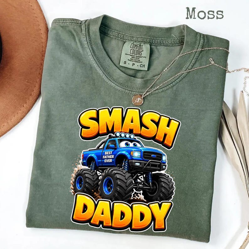 Może przedstawiać: Zielona koszulka w kolorze mchu z napisem "SMASH DADDY" w ż&oacute;łtych i pomarańczowych literach. Niebieska grafika monster trucka znajduje się na środku, z napisem "BEST FATHER EVER". Złoty naszyjnik jest zarzucony na koszulkę.