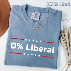 May include: Blue jean-colored t-shirt with the text "0% Liberal" in white, framed by red lines and stars. A gold necklace with a pendant rests on the shirt. The shirt is folded and has a Comfort Colors tag.