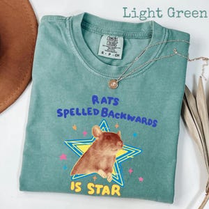 May include: Light green t-shirt with the text "RATS SPELLED BACKWARDS IS STAR" and a graphic of a rat inside a yellow and blue star. The shirt has a Comfort Colors tag and a silver necklace.