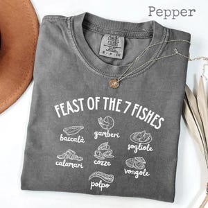 May include: A gray Comfort Colors t-shirt with the text "FEAST OF THE 7 FISHES" and illustrations of seafood. The shirt includes the names of the seafood in white text: baccalà, gamberi, sogliole, calamari, cozze, vongole, and polpo. A silver necklace is draped on the shirt.