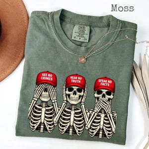 May include: Moss green t-shirt featuring three skeleton figures in the "See No Evil, Hear No Evil, Speak No Evil" pose. Each skeleton wears a red baseball cap with a different phrase: "SEE NO CRIMES", "HEAR NO TRUTH", and "SPEAK NO FACTS".