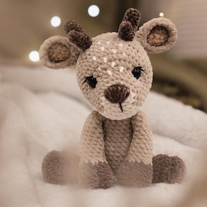 May include: A handmade, plush deer toy in shades of beige and brown. The deer has a crocheted body with a white spotted pattern on its head. It features dark eyes, a brown nose, and small antlers. The toy is sitting on a soft, white surface.